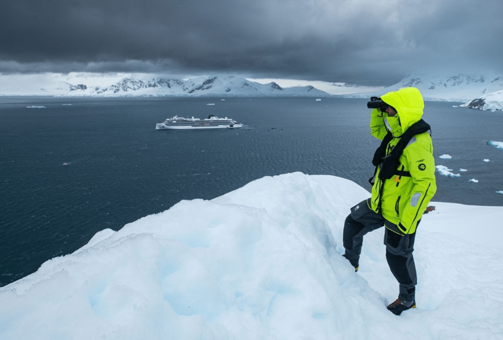 Expedition traveller in yellow jacket surveying icebergs and mountains from a snowy Antarctic ridge with the ship below