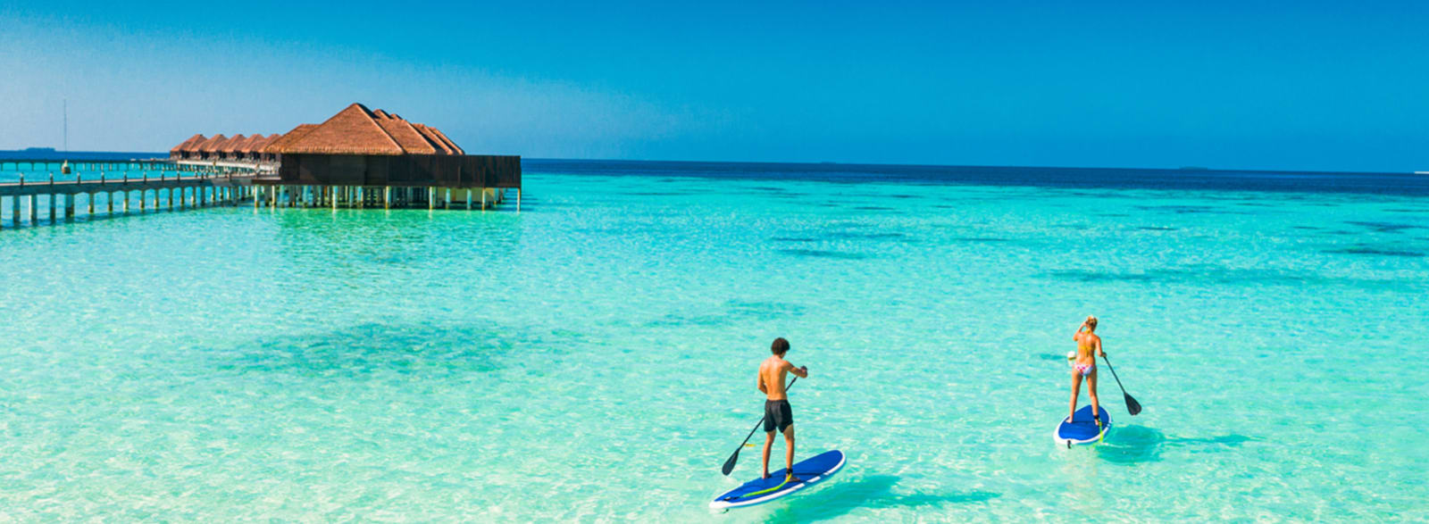 Two people on stand-up paddleboards on a clear ocean, with rows of bungalows in the background
