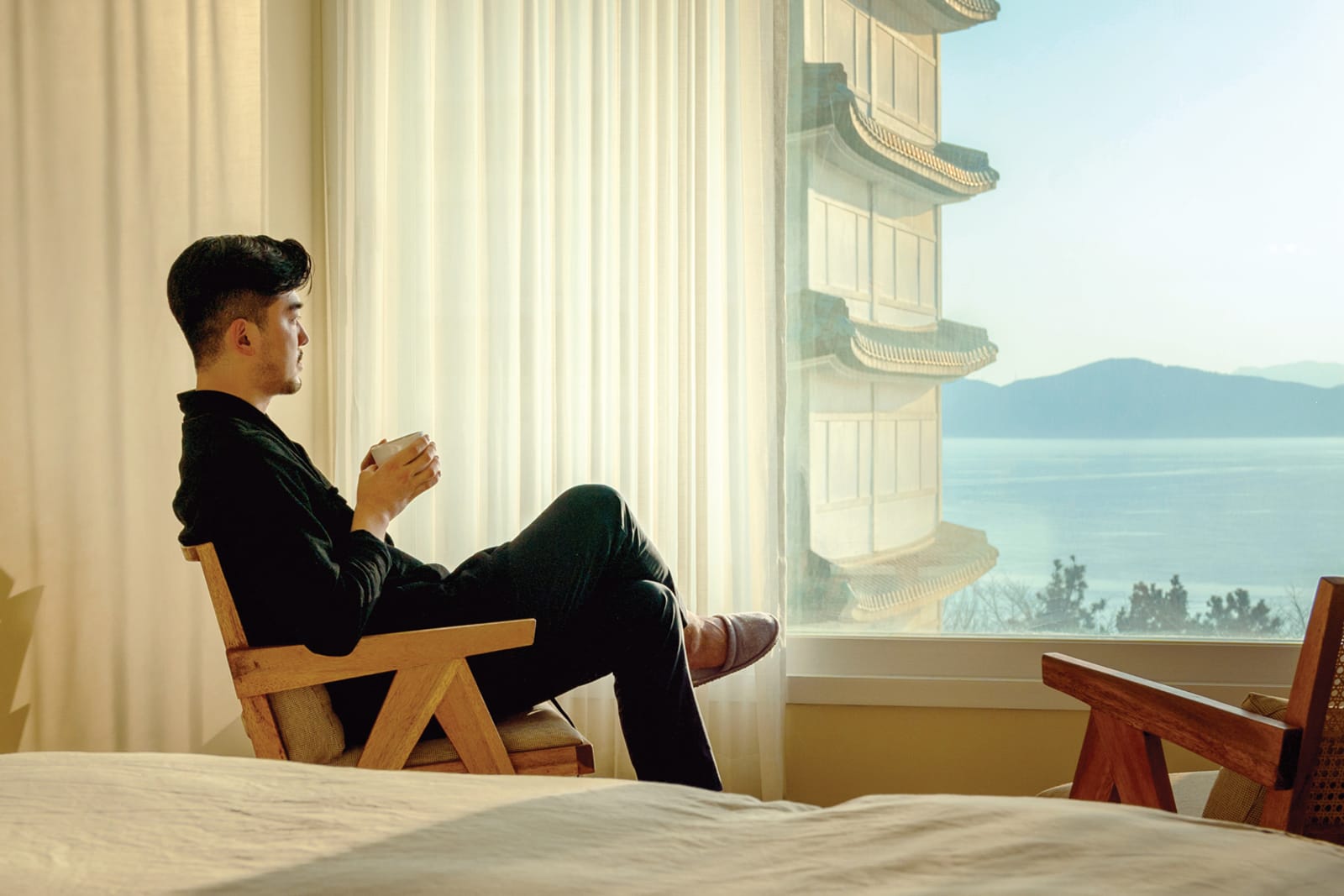 Businessman sitting in a wooden chair by a large hotel room window, holding a cup and looking out at a scenic view.