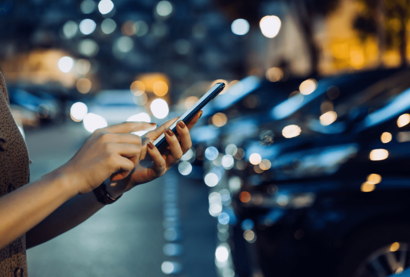 Woman checking her phone on a city street at night surrounded by blurred car lights
