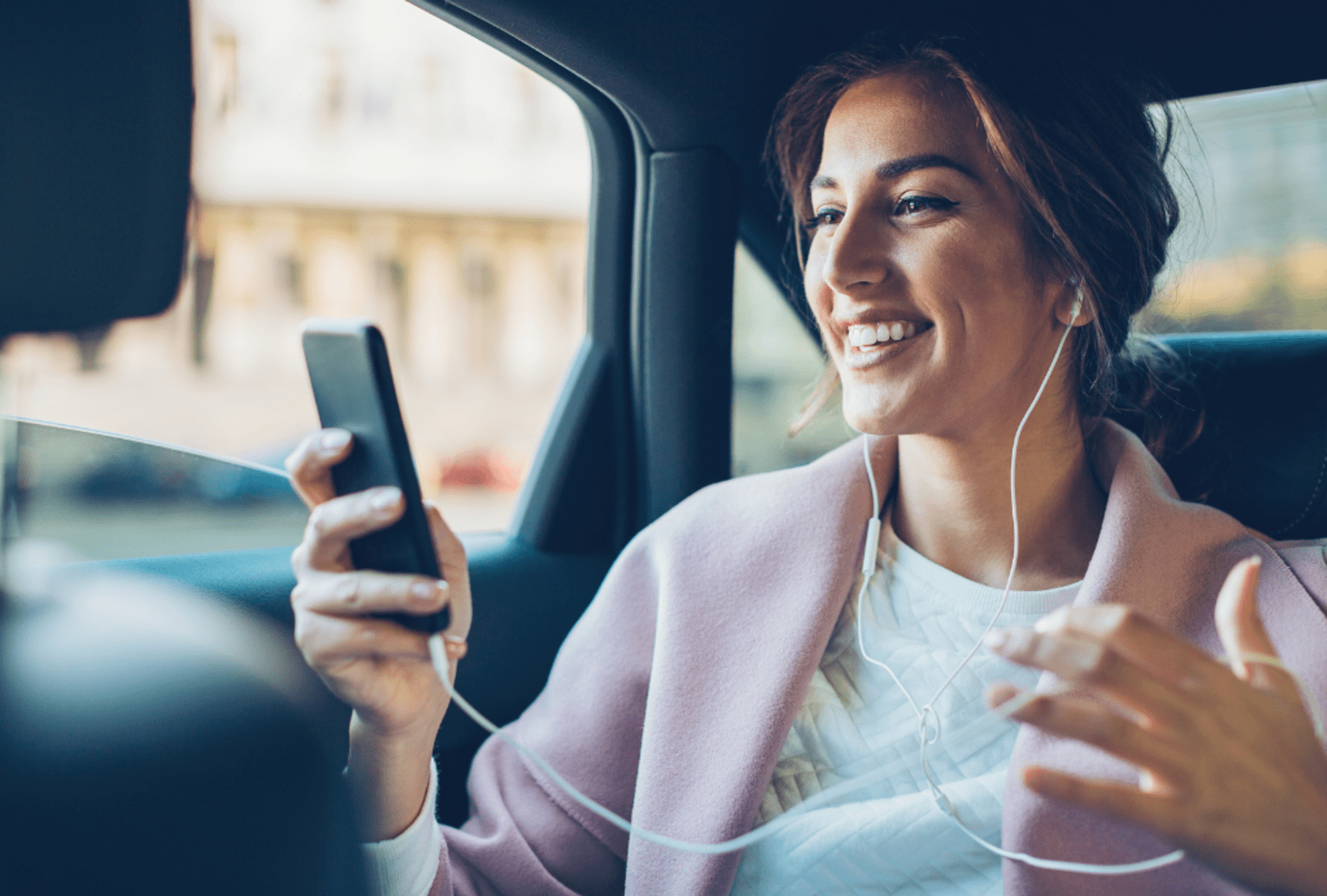 Smiling woman wearing earphones holding a smartphone while riding in the back seat of a car