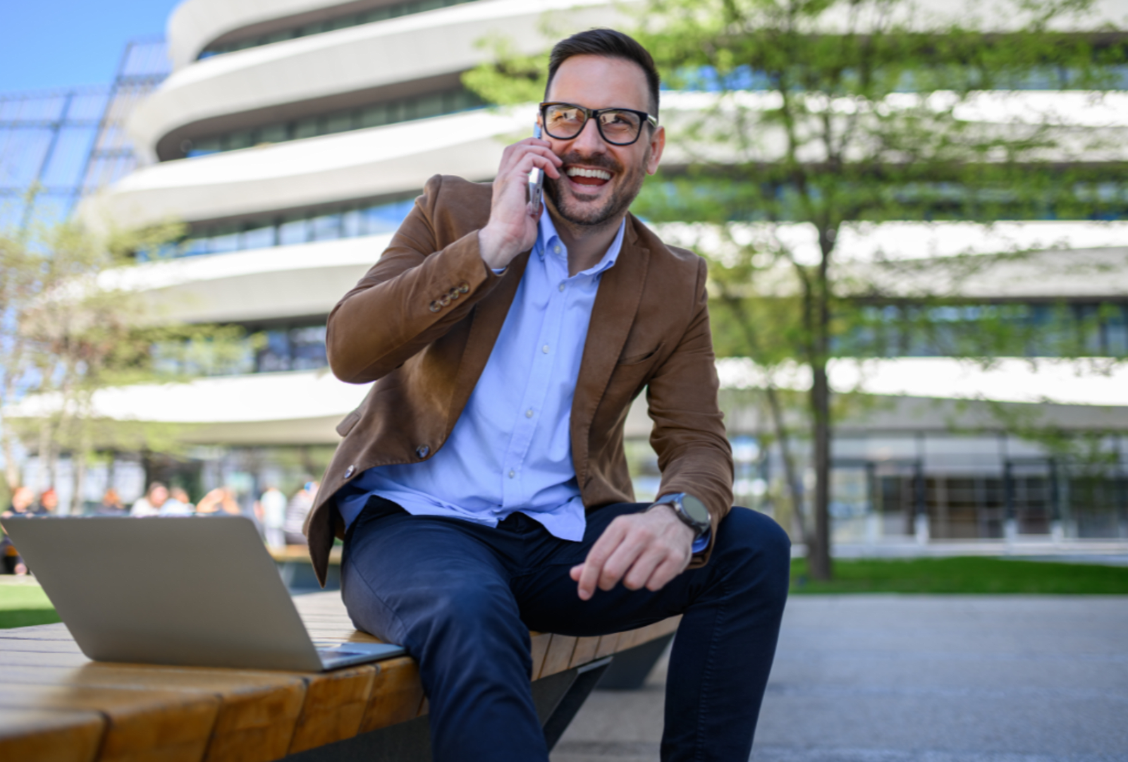 Smiling businessman talking on his phone while sitting outdoors on a bench with a laptop, modern office building in the background