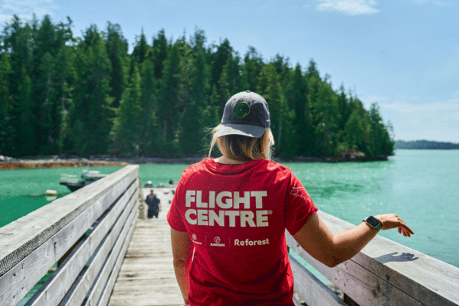 Shot from the back of a long haired person wearing a Flight Centre shirt, walking on a pier out to a lake