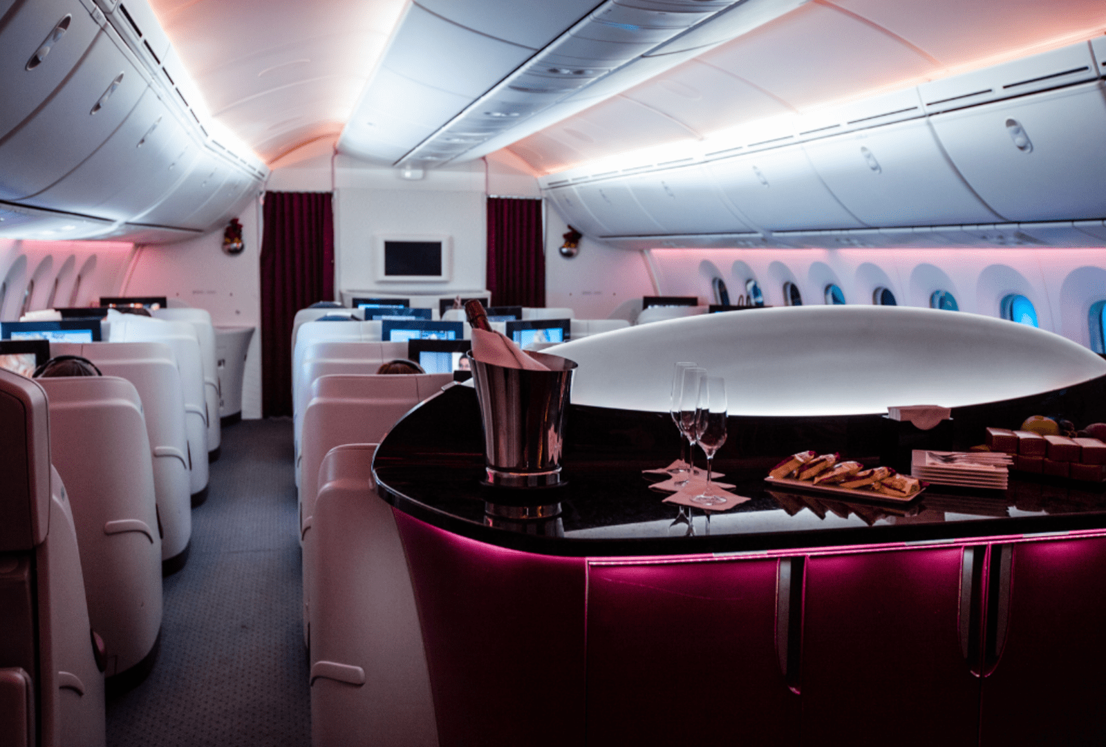 The luxurious interior of a Qatar Airways business class cabin, featuring a curved bar with champagne on ice, wine glasses and snacks, surrounded by private white pod seats with mood lighting