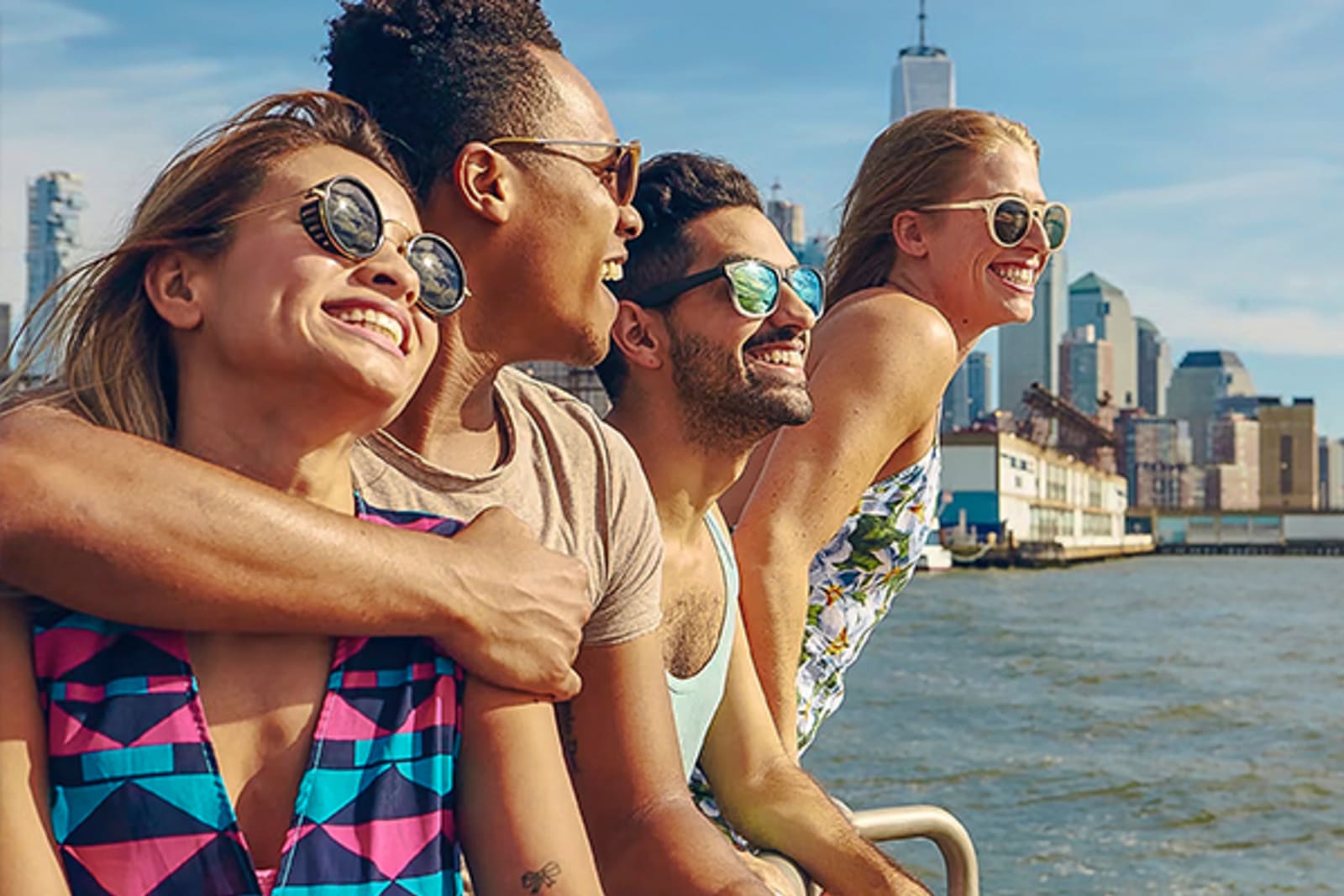 Four people smiling, wearing sunglasses sitting on the edge of the water in a city