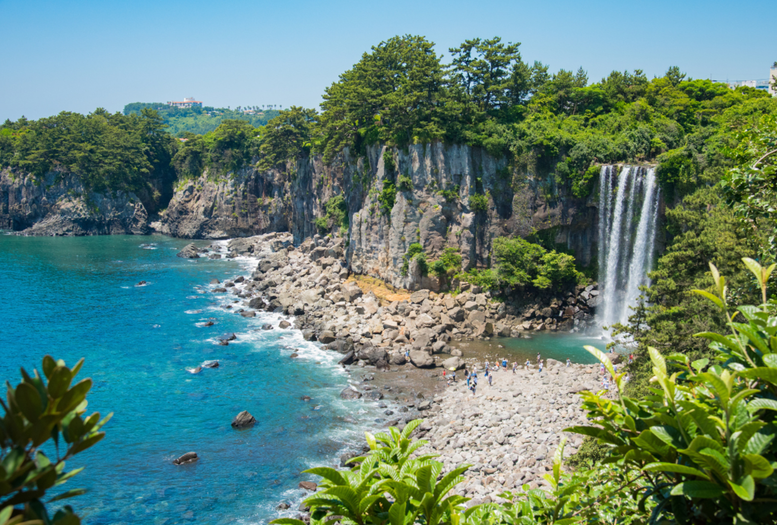 A dramatic coastal cliff face with a waterfall cascading onto a rocky shoreline on Jeju Island, South Korea, with turquoise waters and visitors exploring the beach below