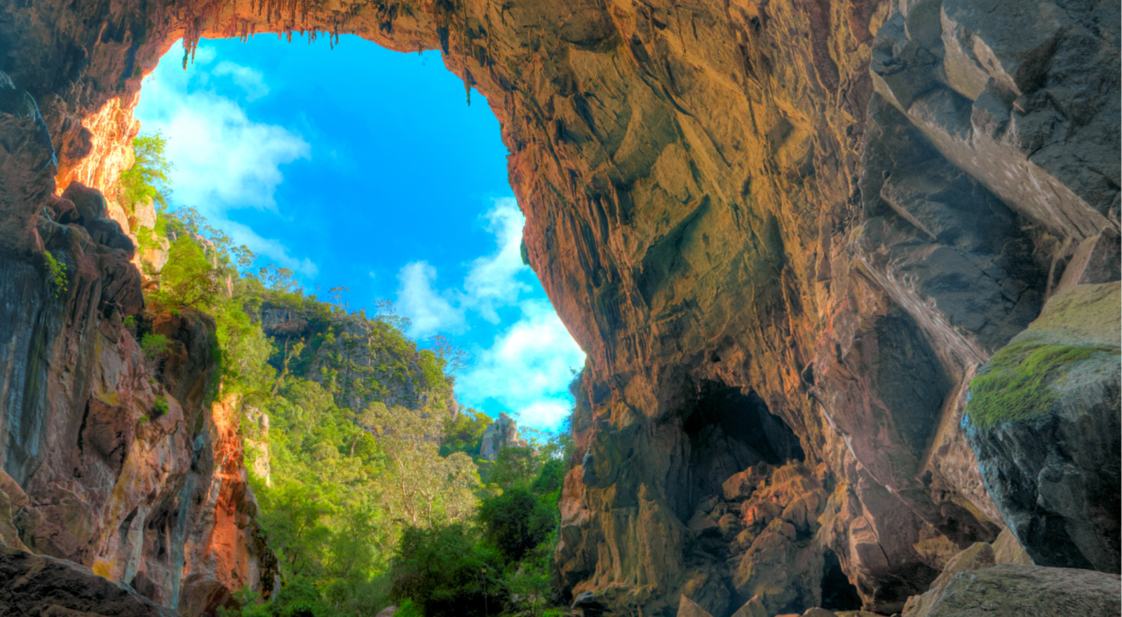 A sunlit rock cave archway at Jenolan Caves framing a lush green rainforest valley and bright blue sky in the distance