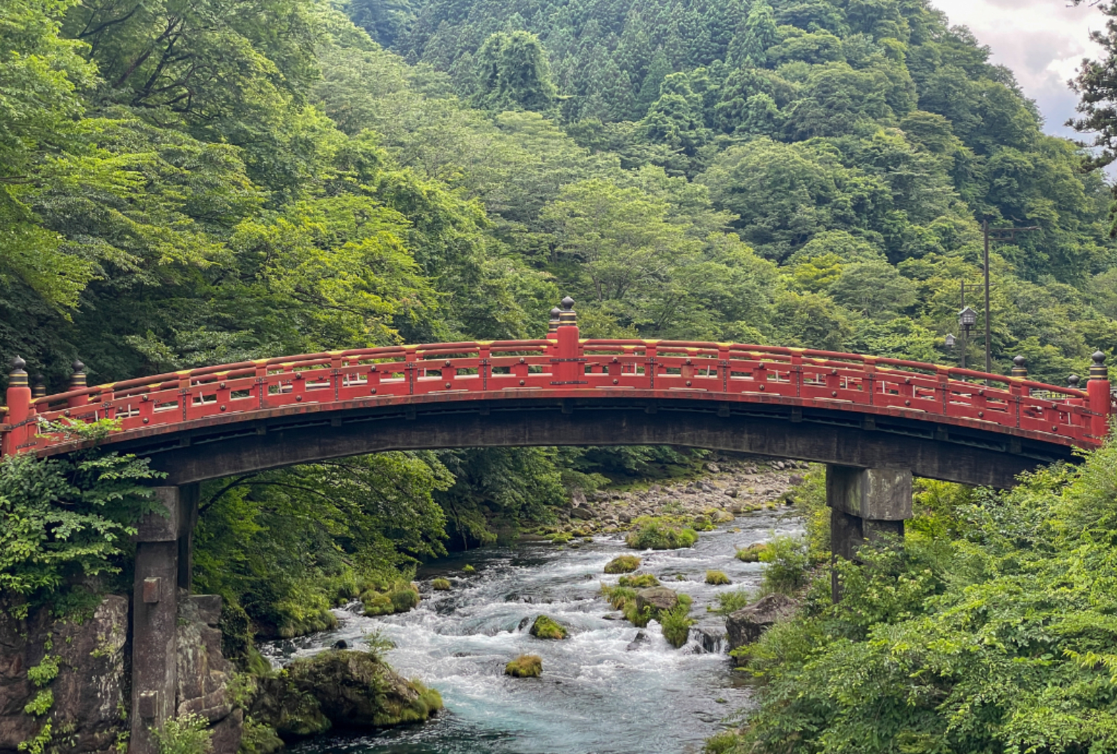 The iconic red Shinkyo Bridge arching over a rushing river surrounded by lush green forest in Nikko, Japan