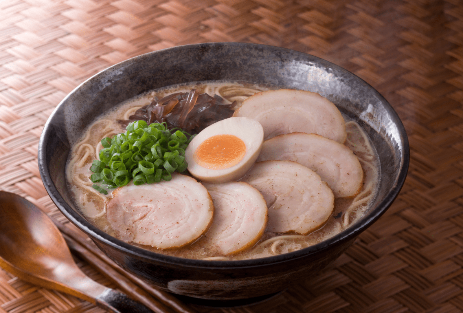 A bowl of Japanese ramen topped with chashu pork slices, a soft-boiled egg, spring onions and mushrooms, served with chopsticks and a wooden spoon