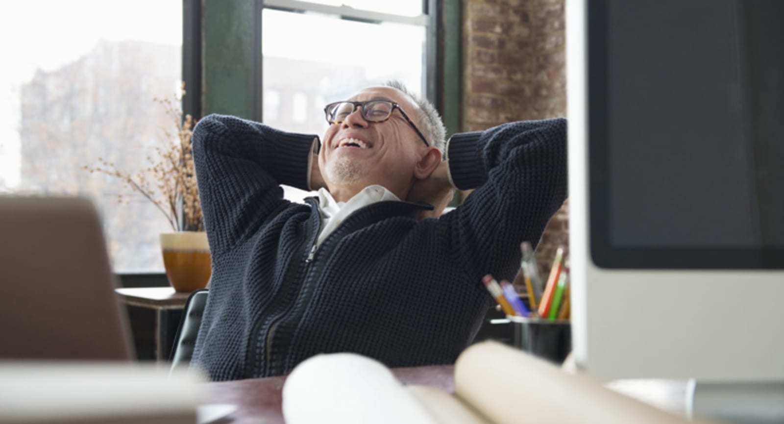 Elderly businessman relaxing in a chair in front of a desk covered in papers, pencils and a computer monitor