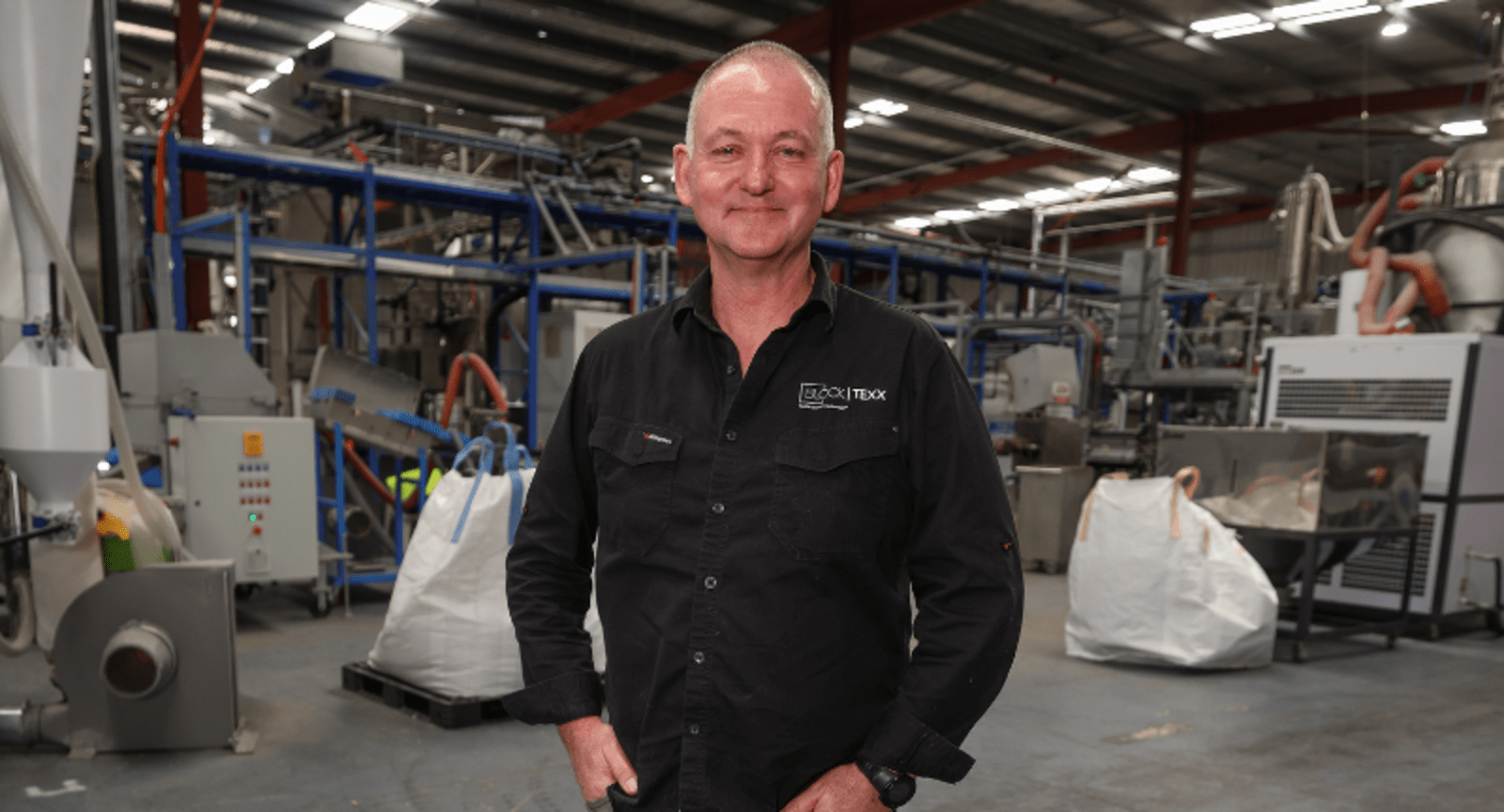 Balding businessman in a black button-up shirt standing in a warehouse filled with equipment