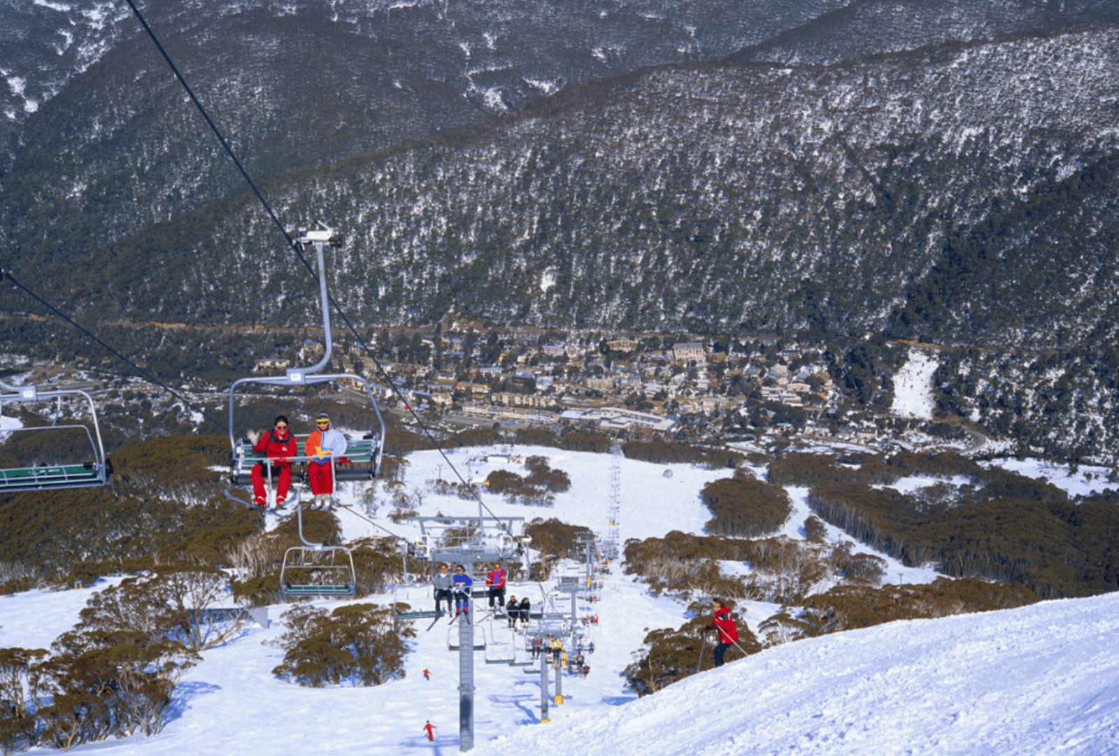 Skiers riding a chairlift above snow-covered Thredbo slopes with the alpine village and mountain ranges stretching into the distance below
