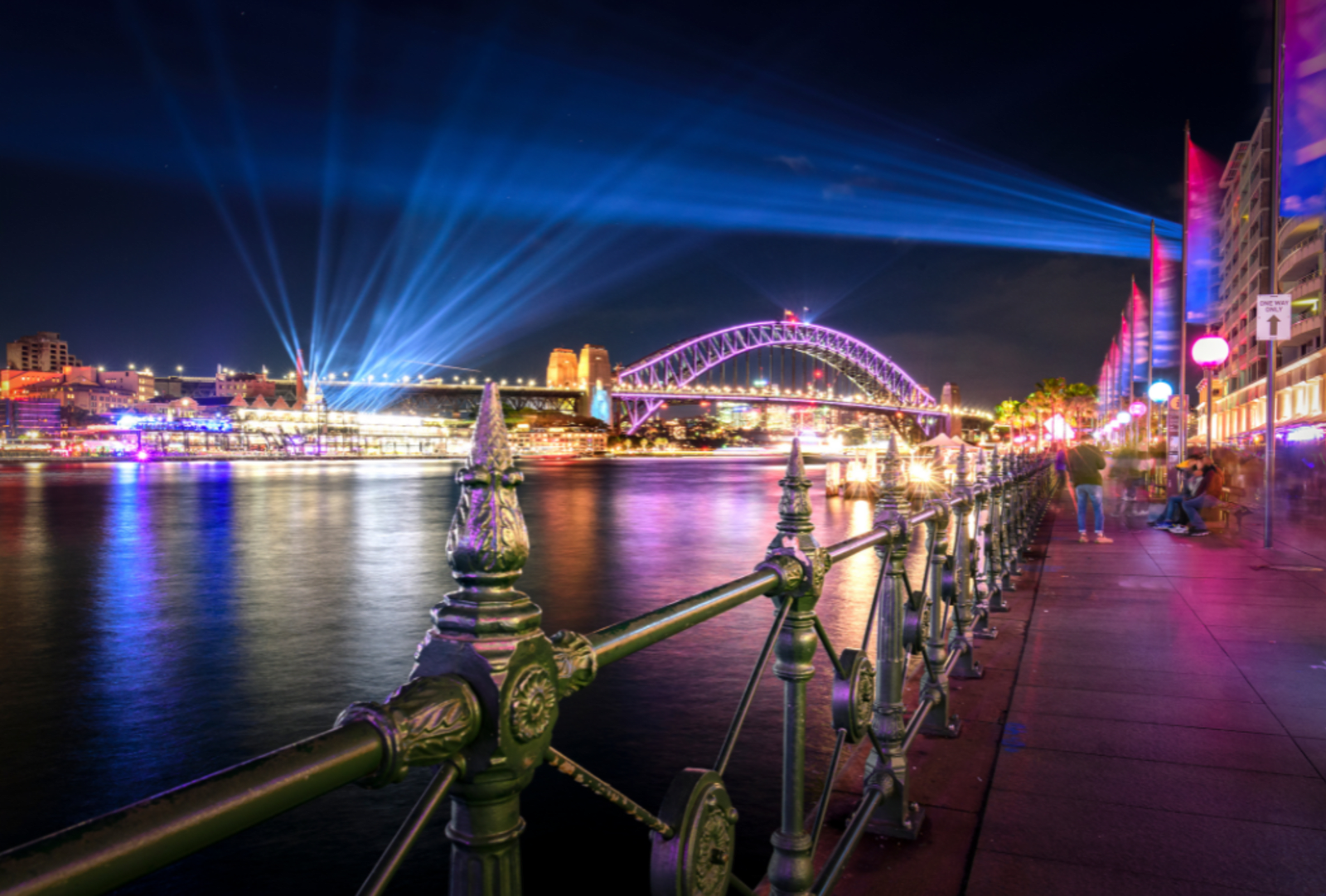 Sydney Harbour Bridge lit in violet with dramatic blue light beams radiating across the harbour waterfront during Vivid Sydney at night