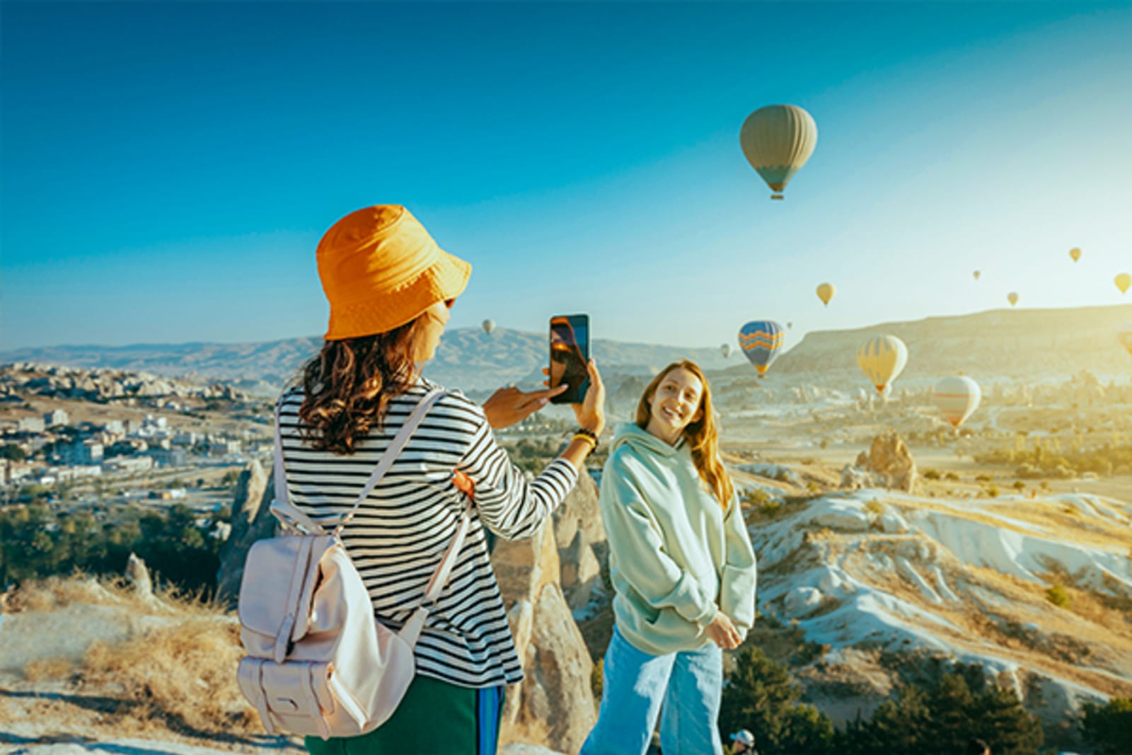 Two women taking pictures with multiple hot air balloons in the background