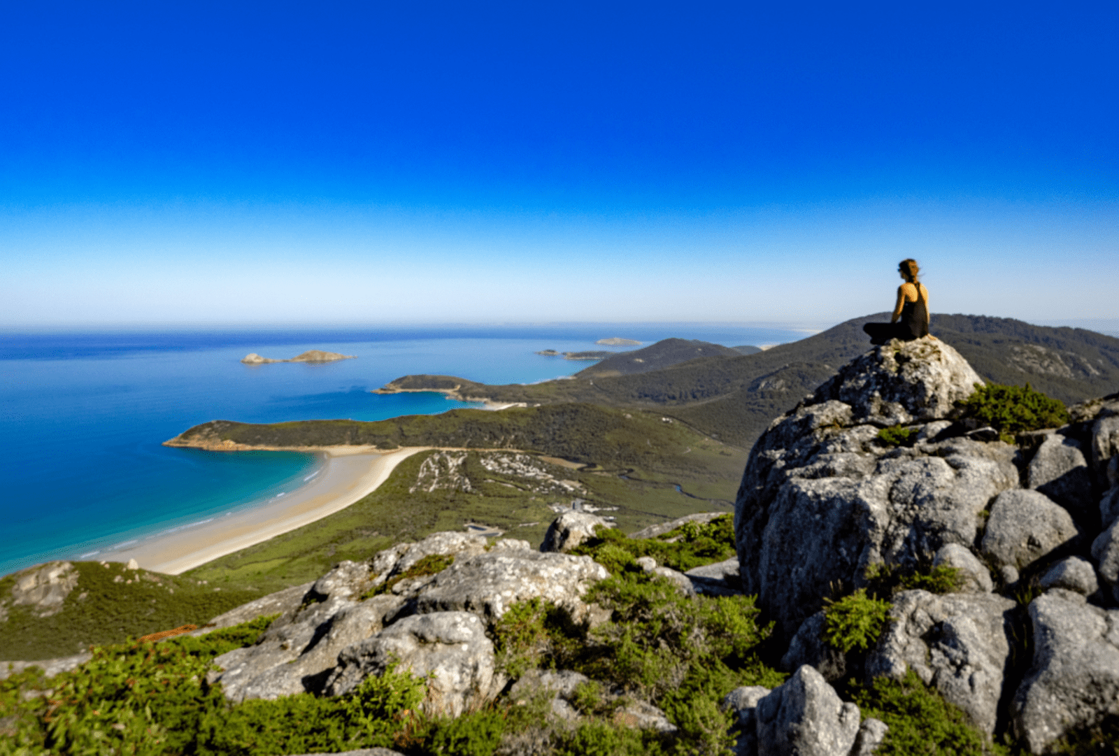 alternative text. Woman sitting on a granite rock summit at Wilsons Promontory gazing over sweeping coastal bays turquoise waters white sand beaches and green headlands
