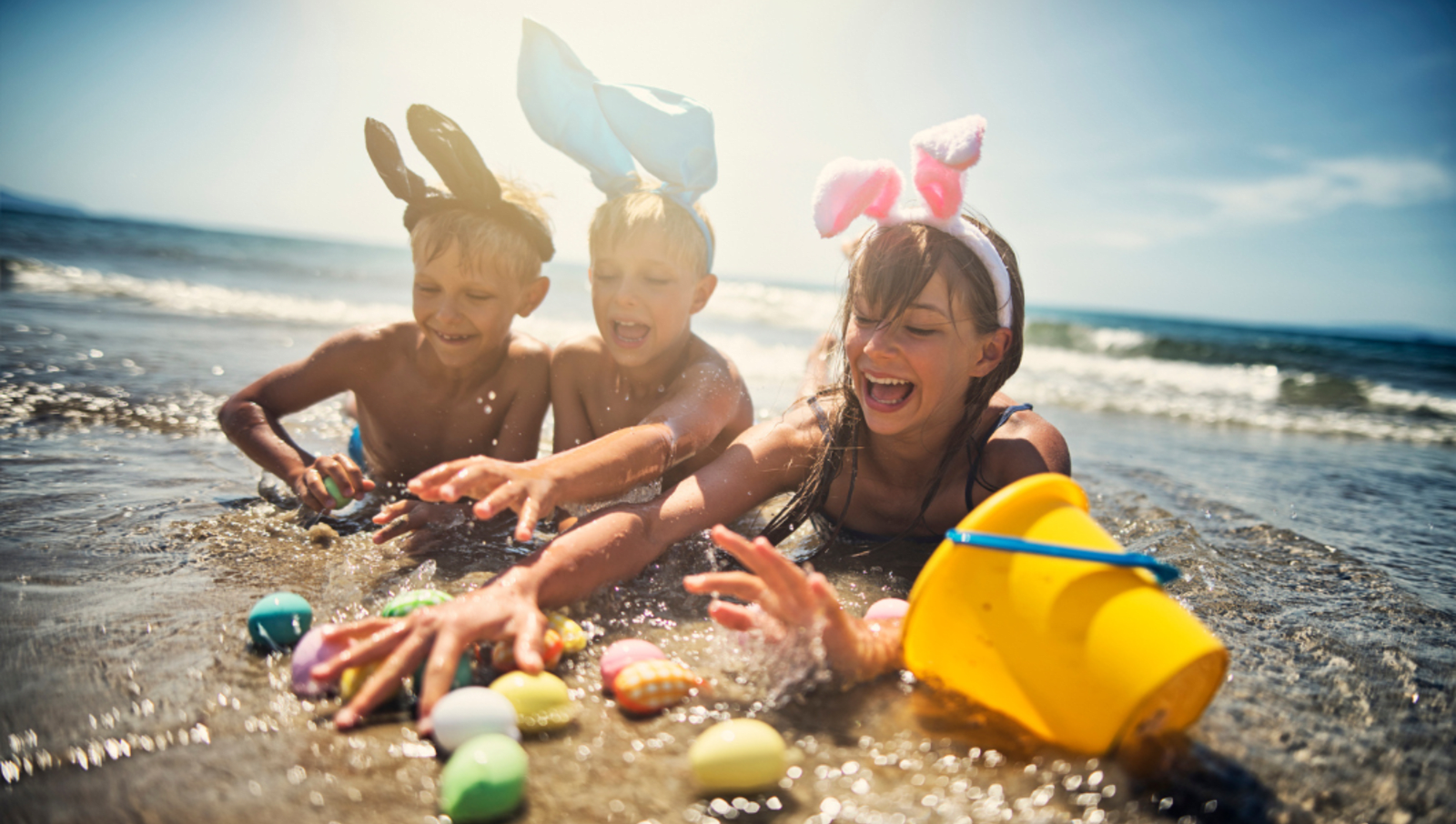 kids wearing bunny ears play in ocean with yellow bucket and coloured Easter eggs 