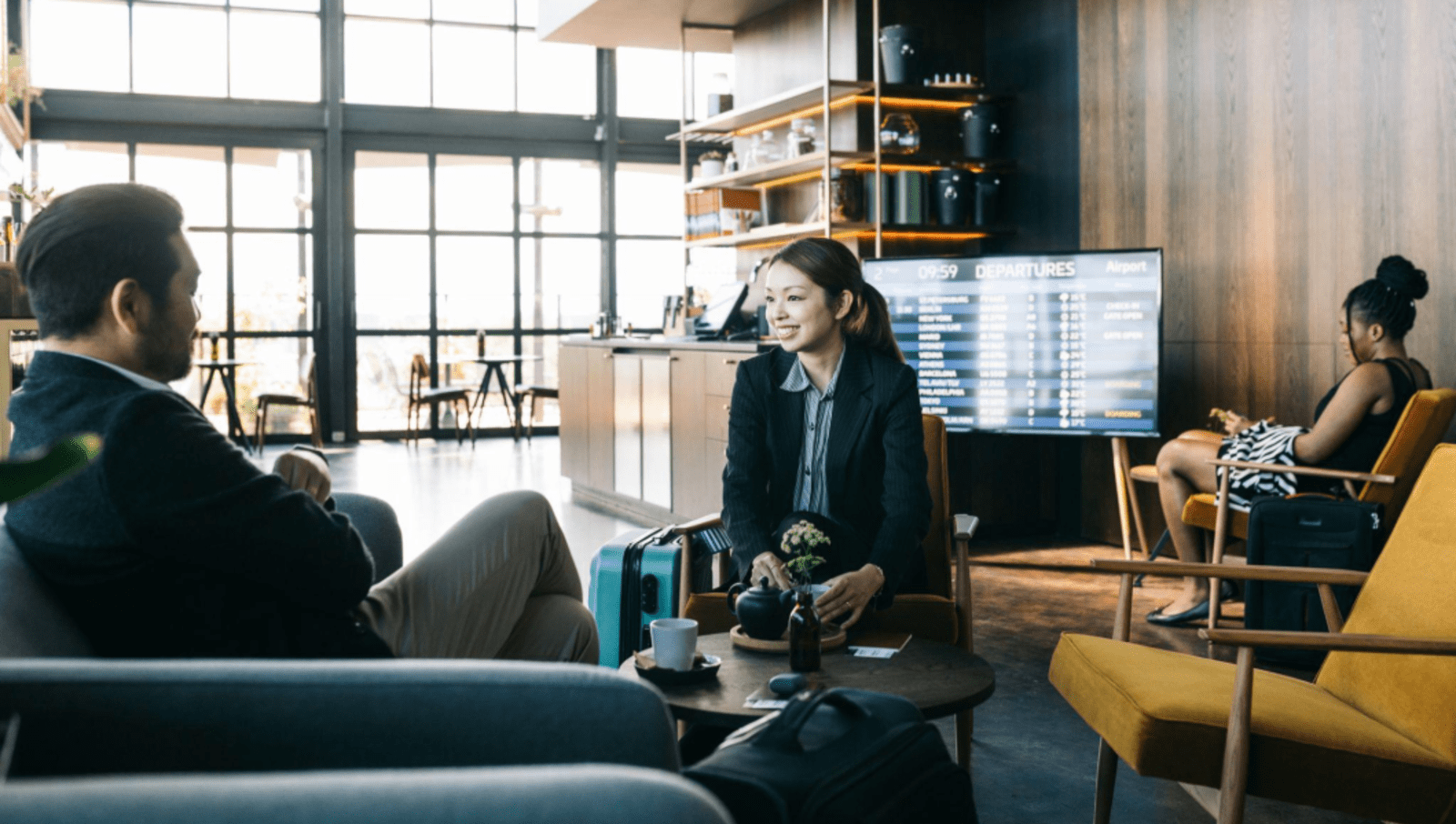 two people in business clothing sit in an airport lounge with coffee on a table in front of them and another person in the background