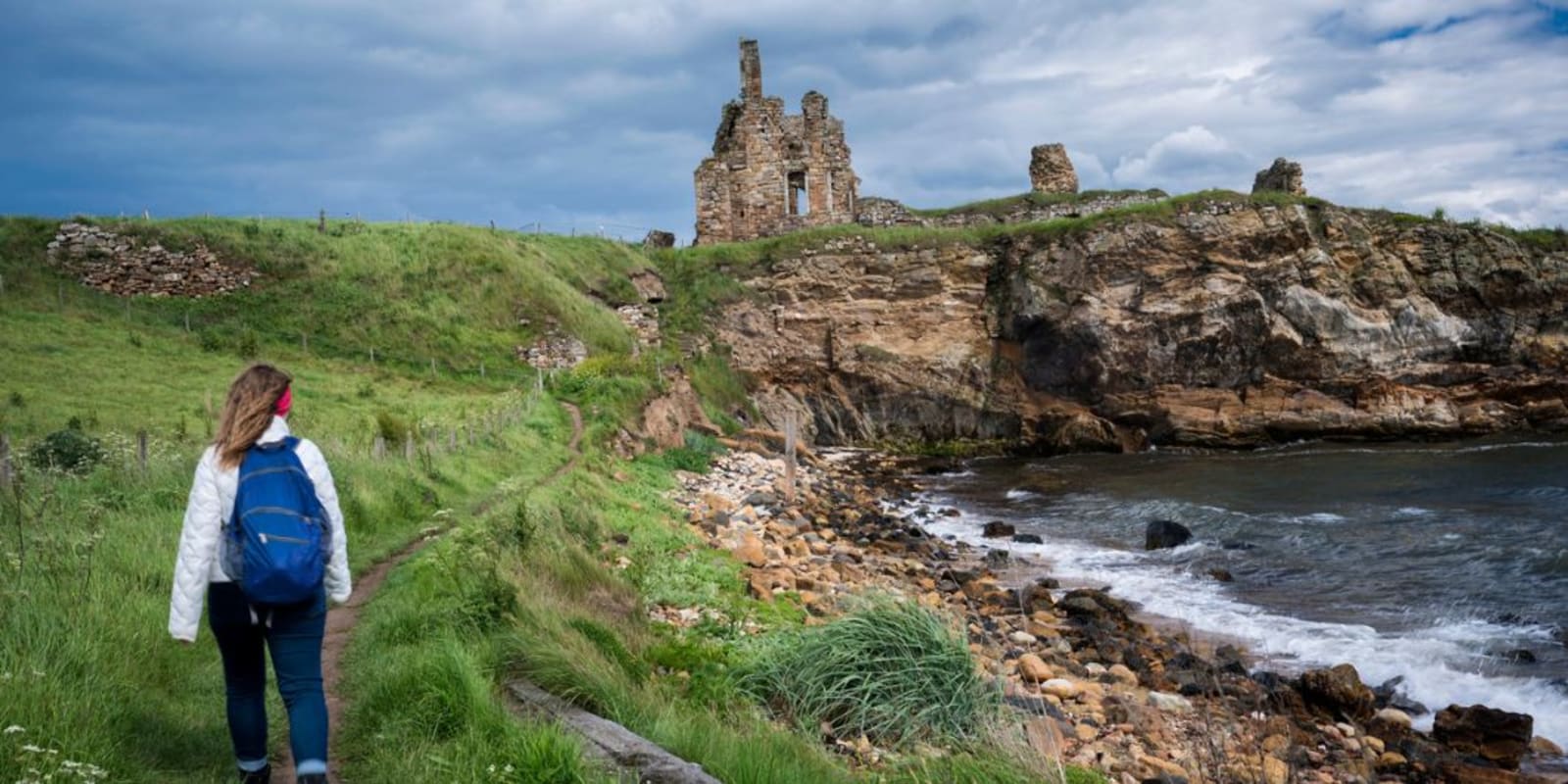 Blond female hiker walks toward the ancient stone ruins of Newark Castle on the rocky coast of the North Sea along the Fife Coastal Path north of Elie, Kingdom of Fife, Scotland, UK, Europe