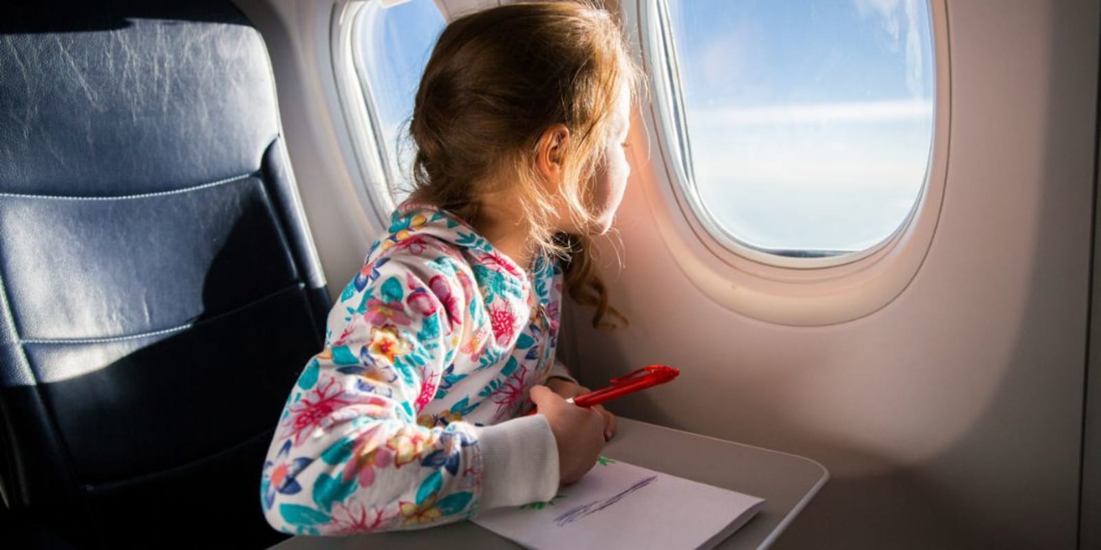 A young kid on a plane looking at the window with a colouring book in front of her.