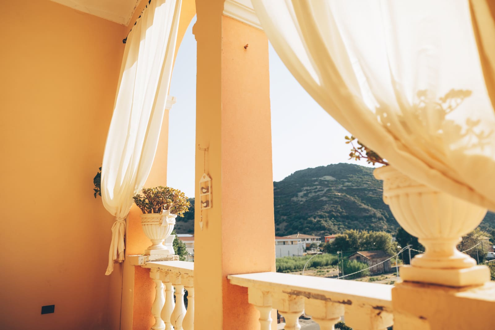 Looking out from a balcony. The balcony is a tan colour with curtains blowing in the wind. Over the balcony, stunning green mountains can be seen.