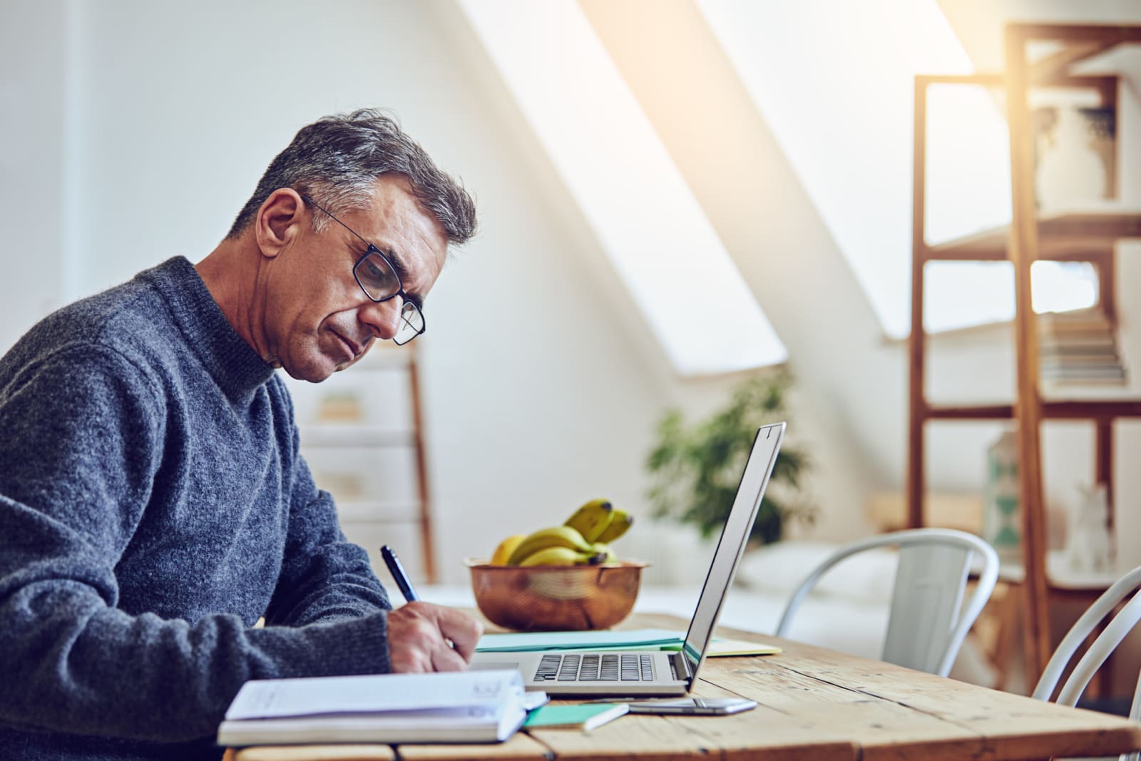 person sits at table, writing in notepad with a laptop in front of them