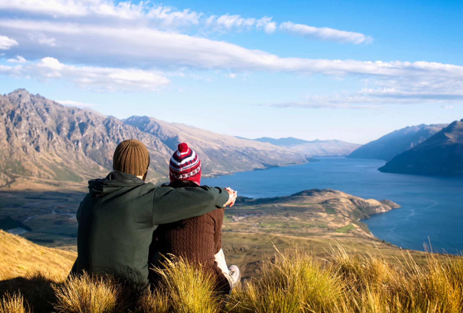 A couple sits on a tussocky hilltop overlooking a serene lake