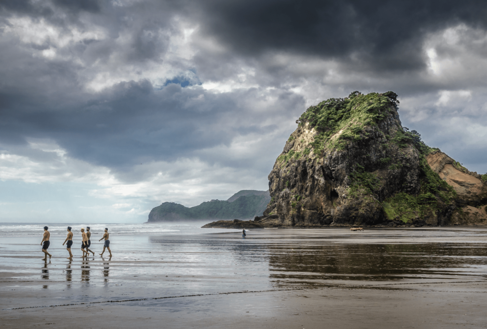 Swimmers wander across the black sands of Piha Beach at low tide with Lion Rock in the background