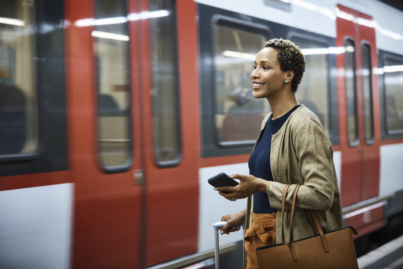 person stands with suitcase, bag and phone waiting to catch train