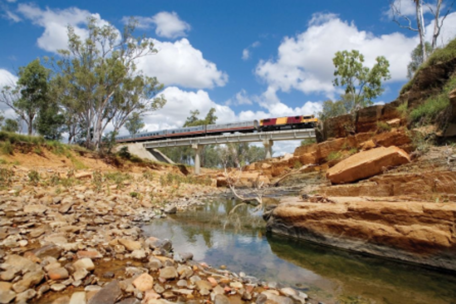 spirit of the outback train travelling along train track above small river surrounded by rocks