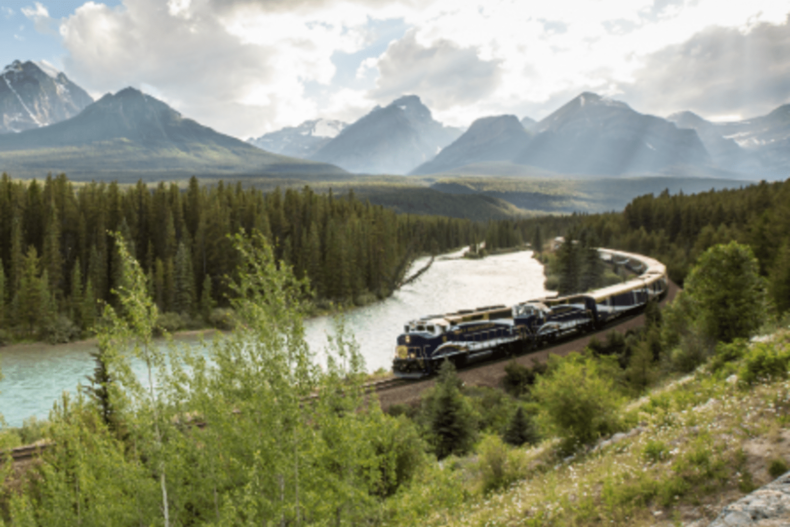 rocky mountaineer train running along traintrack surrounded by greenery and water with mountains in the background