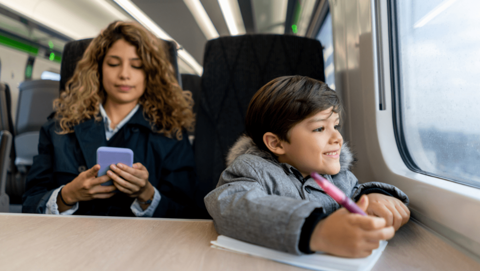 Young boy smiling and looking out the window of a train as he holds pen to paper and his mum sits beside him on her phone