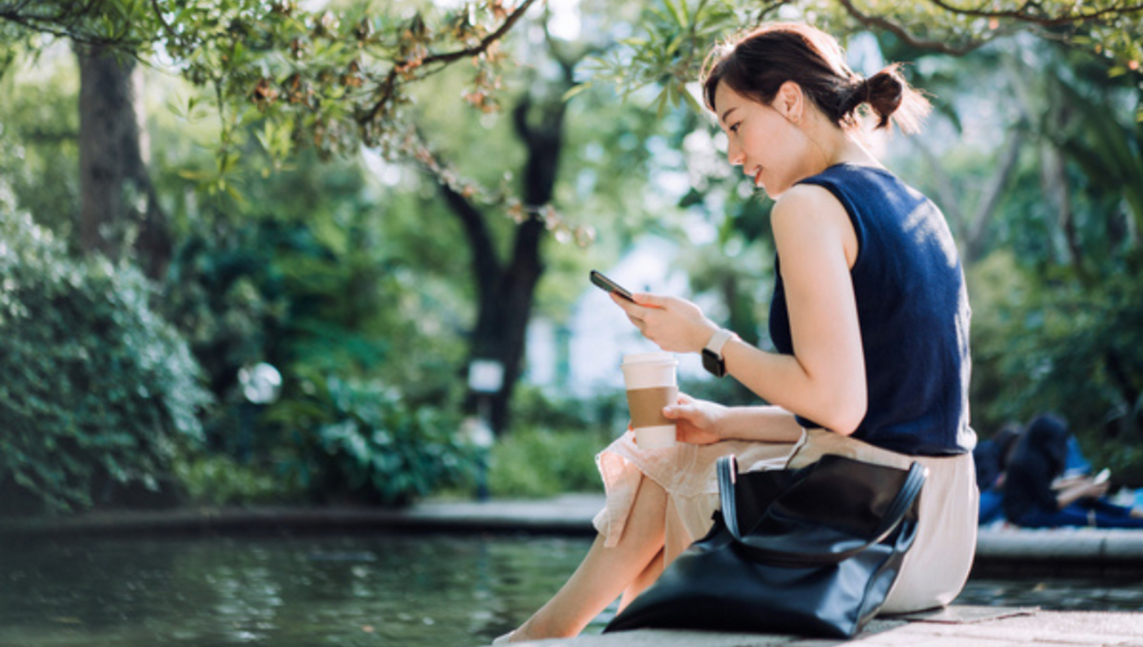 Person in smart casual clothes sits with their feet in a pond, coffee in their and and looking at phone