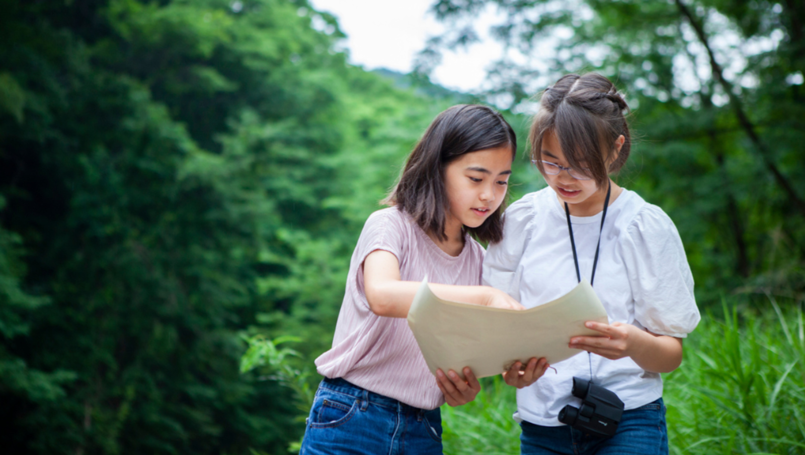 Two girls looking and pointing at a map they are holding while standing in a lush green jungle