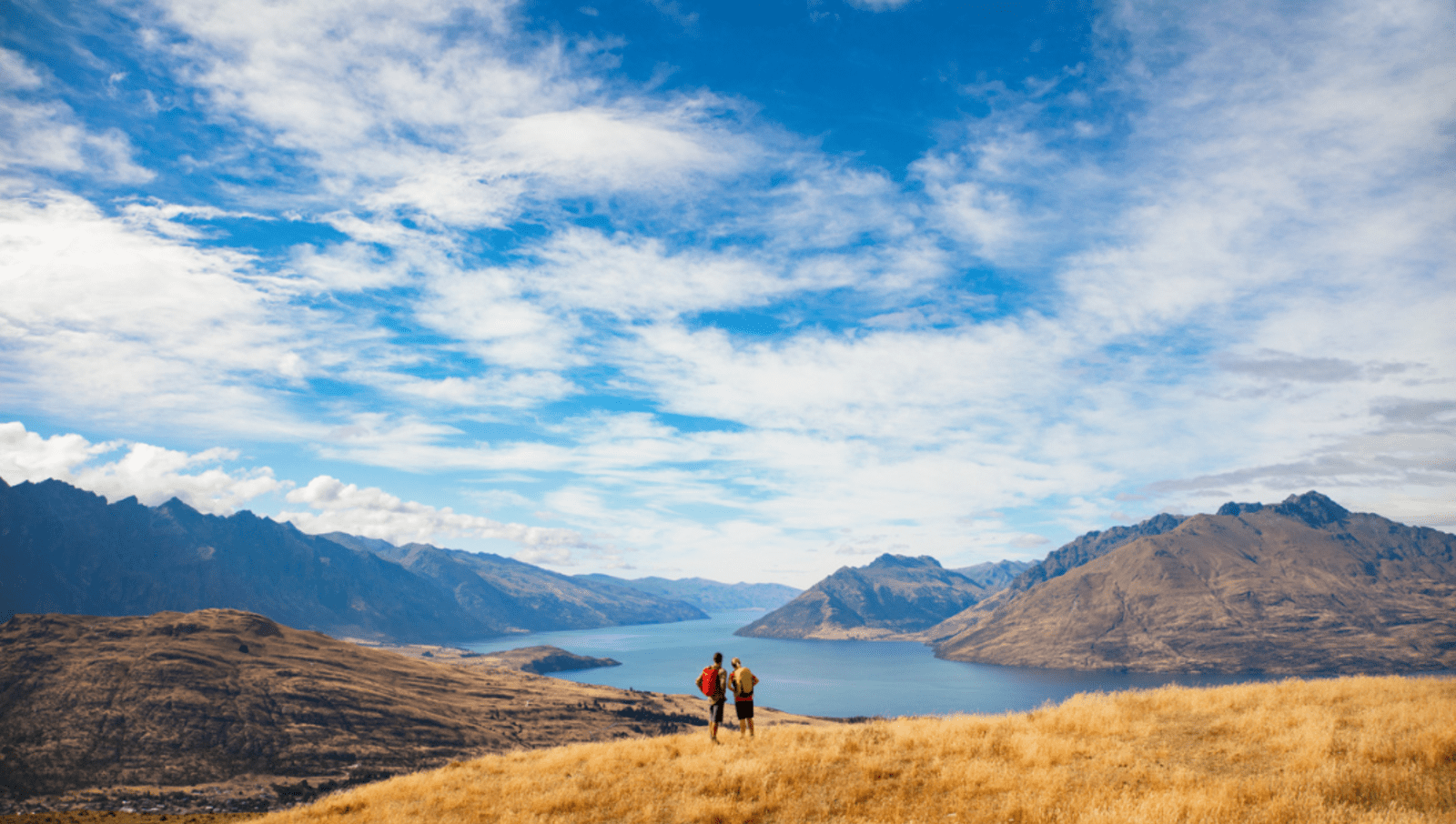 two men with backpacks standing on mountain overlooking more mountains surrounding a lake
