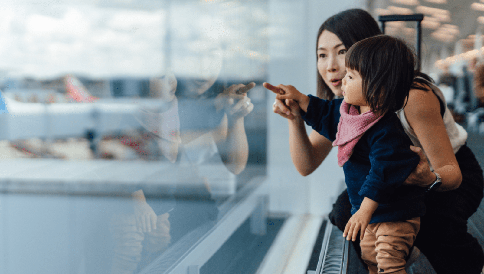 Mum and toddler pointing out the window of an airport and looking at the planes