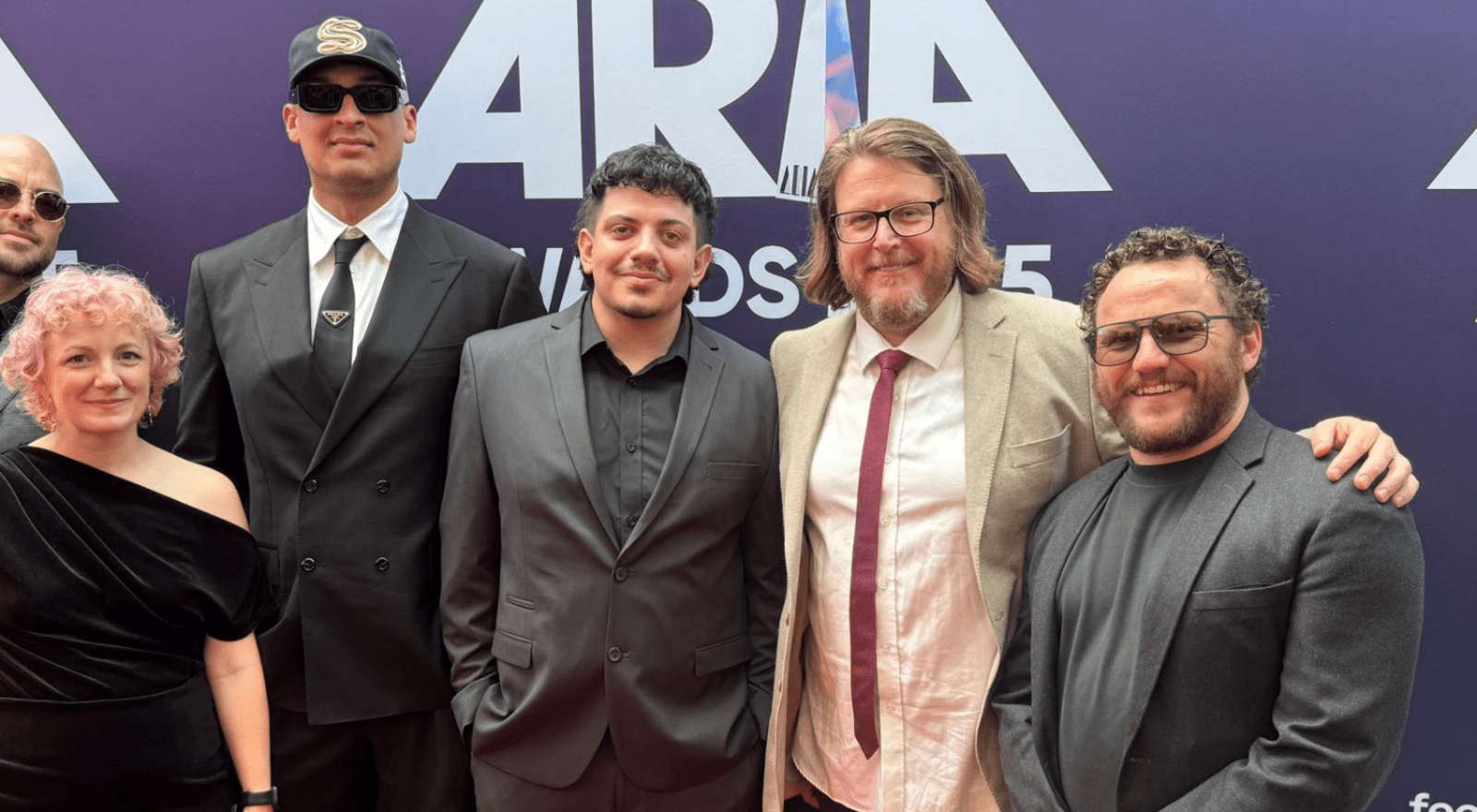A group of six people posing together on the ARIA Awards red carpet in front of a purple ARIA Awards branded backdrop. From left to right: a person partially visible at the back left wearing sunglasses; a woman with short pink curly hair in a black one-shoulder dress; a tall man in a black suit, white shirt, tie, and green cap with sunglasses; a man in a dark grey suit with a black shirt; a man with long blonde-grey hair and a beard wearing a beige blazer, light shirt, and burgundy tie; and a man with curly