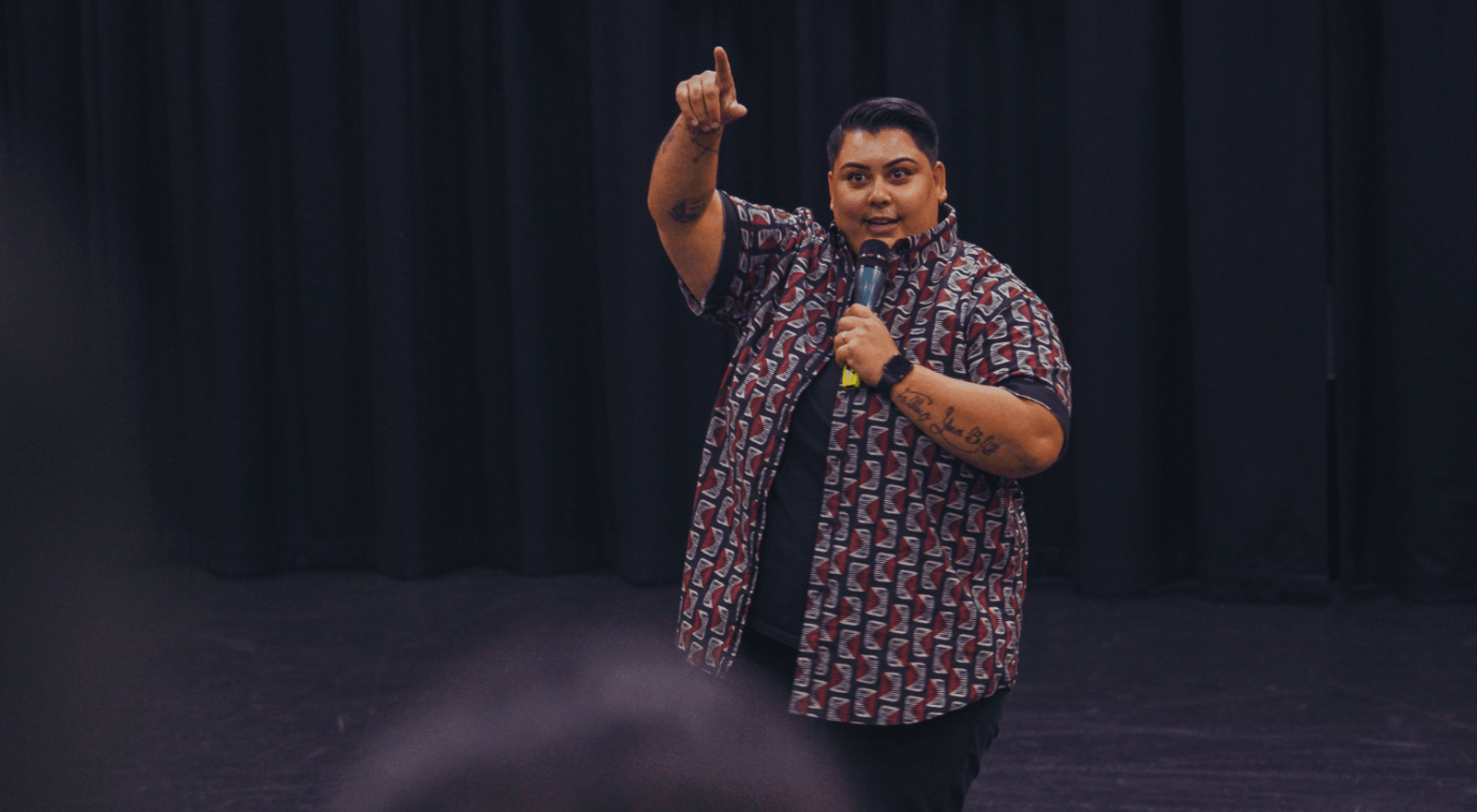 A performer stands on a darkened stage holding a microphone in one hand while pointing upward with their index finger. They are wearing a short-sleeved, open button-up shirt with a bold red, white, and black geometric pattern over a black t-shirt, black pants, and a black watch. They have short dark hair, visible tattoos on both forearms, and are mid-performance with an engaged, expressive facial expression. A dark curtain forms the backdrop, and a blurred audience is partially visible in the foreground.