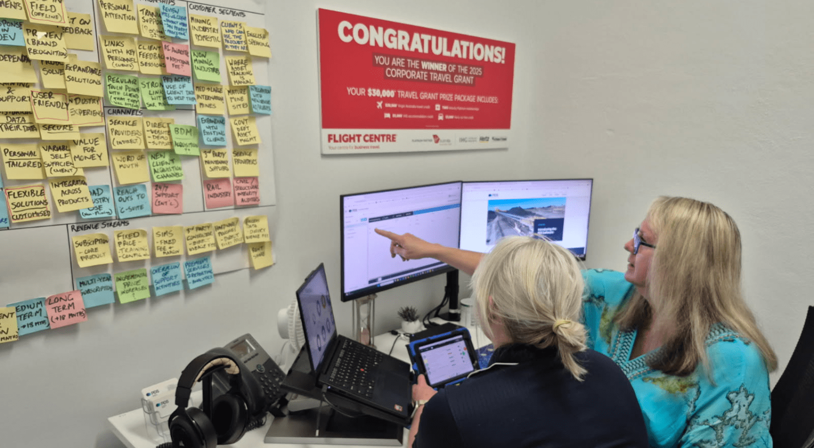 Two women with blonde hair sit together at a desk in an office, looking at a dual-monitor computer setup. One woman, wearing a teal patterned top and glasses, points at one of the screens while the other, in a dark top, holds a tablet. The desk also has a laptop, a desk phone, and headphones. On the wall behind them hangs a red Flight Centre "Congratulations! You are the winner of the 2025 Corporate Travel Grant" poster advertising a $30,000 travel grant prize package. To the left, a large whiteboard wall i