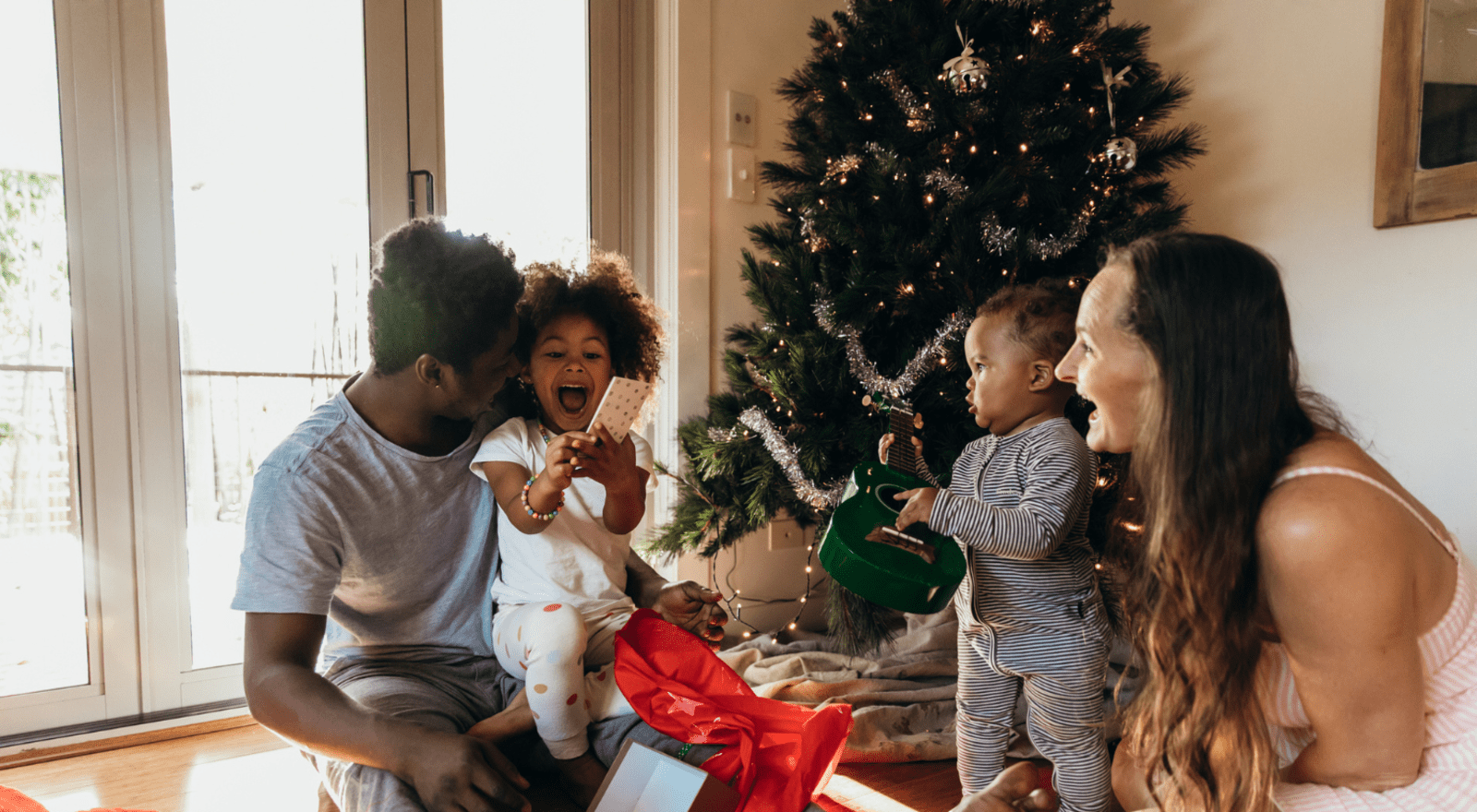 family of four sitting on ground by christmas tree unwrapping presents