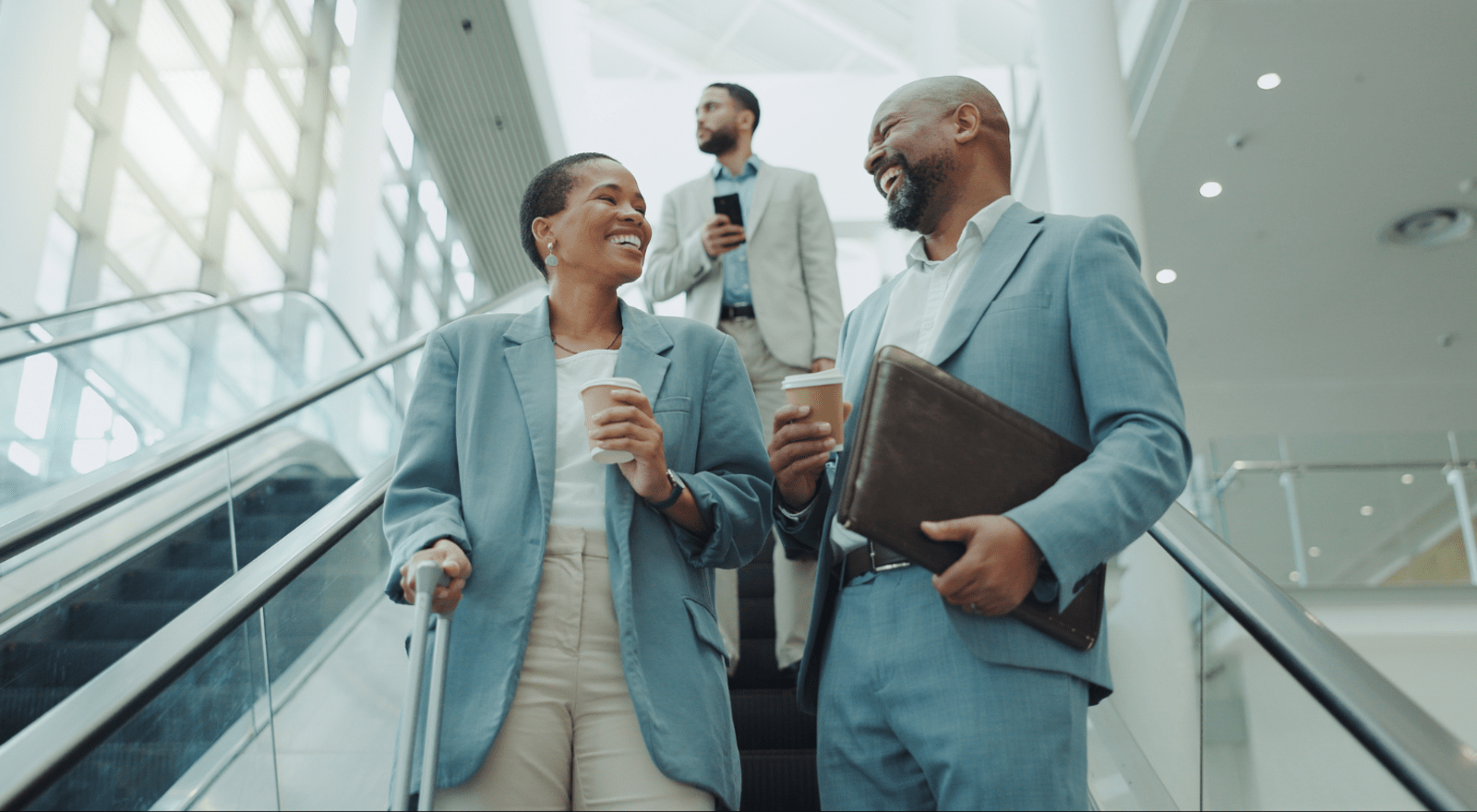 Two business people on an escalator holding coffee and laughing at each other