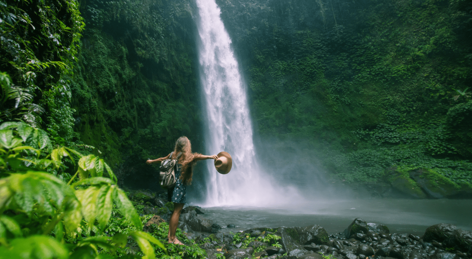 woman standing at base of waterfall with arms out holding hat in indonesia 
