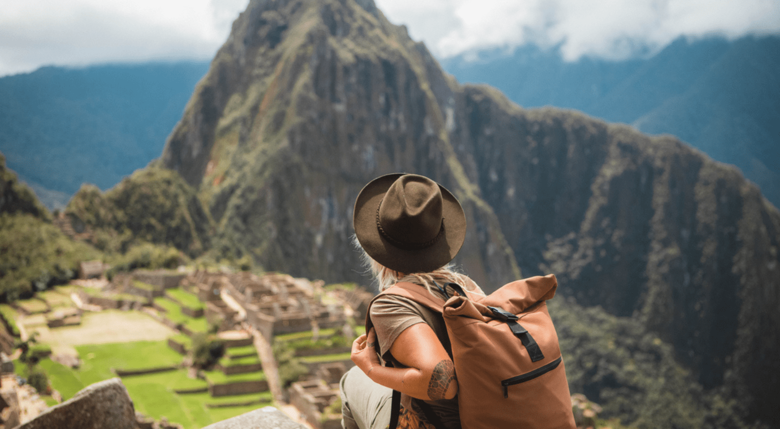 woman wearing hat and backpack looking over macchu picchu from cliff