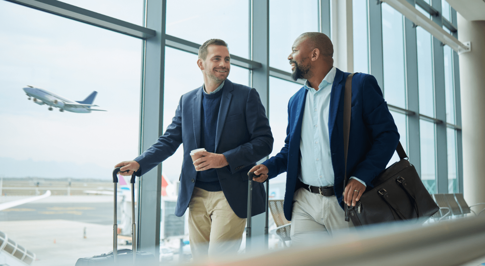 two business men smiling and looking at each other while walking through an airport with plane in the background