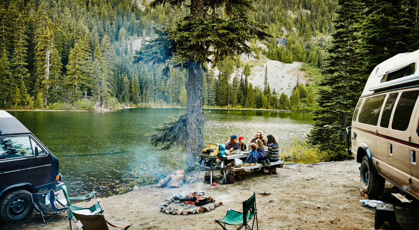 Group of adults and children sitting at picnic table by lake with fire nearby