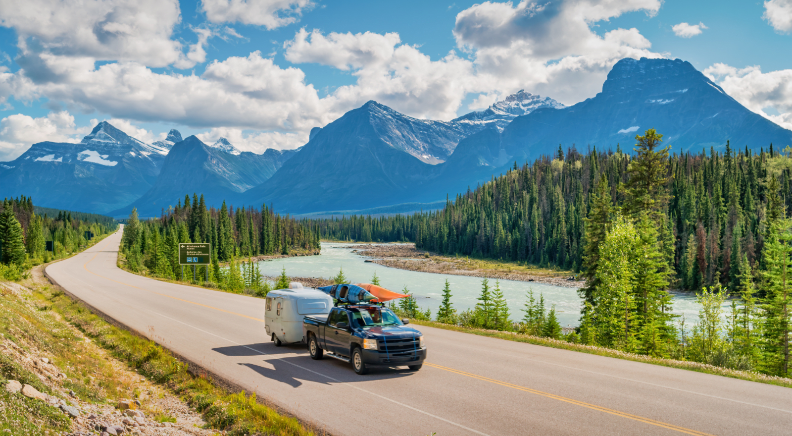 truck pulling campervan along road in canada with mountains and lake in background