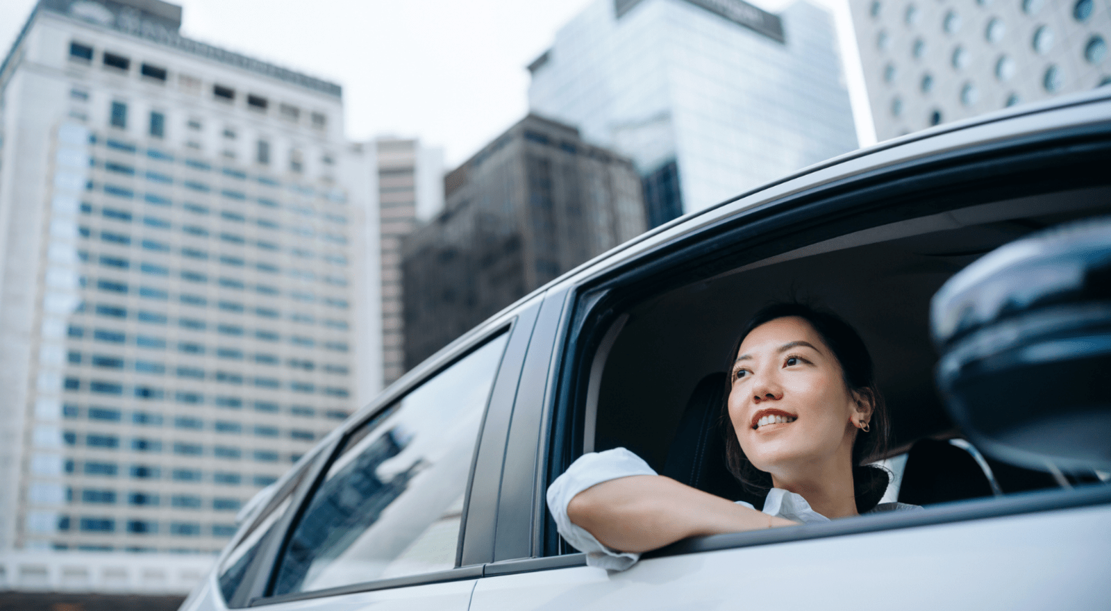A girl in business atire sitting in a parked car looking at the window. The car is parked in a city.