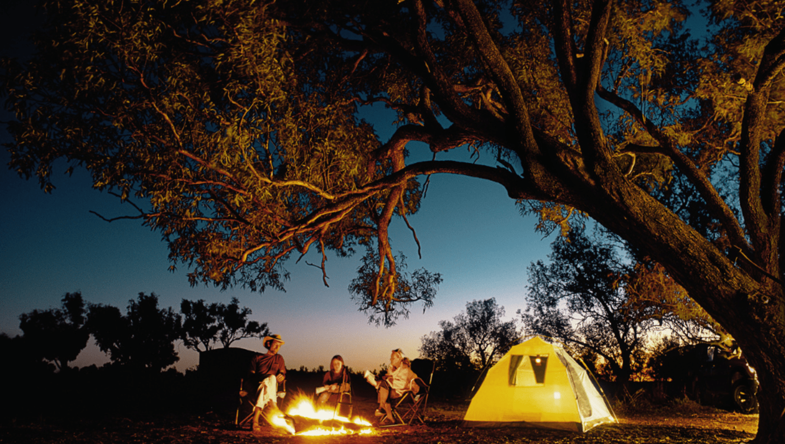 family of two adults and a pre-teen child sit by a camp fire under a big tree with yellow tent next to them 