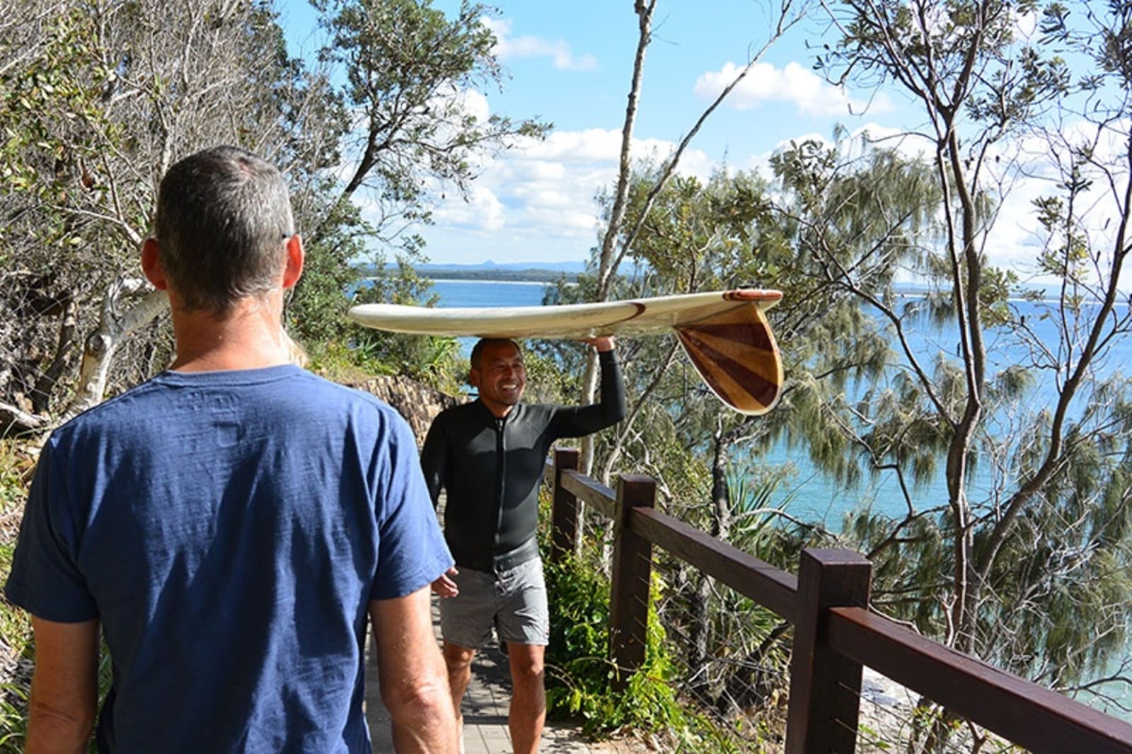 Two people walking past each other on a trail near a beach. One man has a surf board on his head and is smiling.