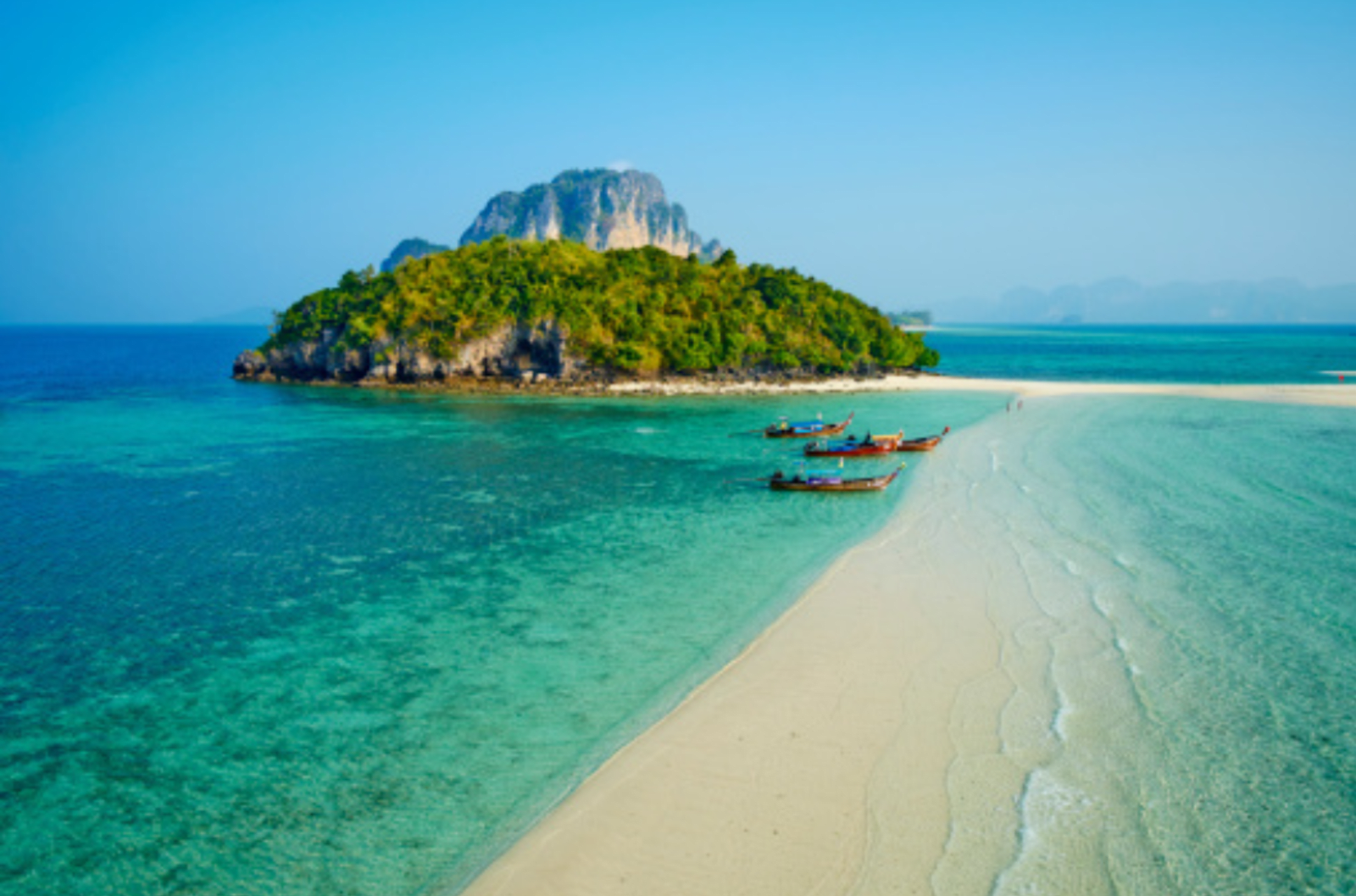 Clear blue waters with a strip of sand running through the middle and greenery in the background.