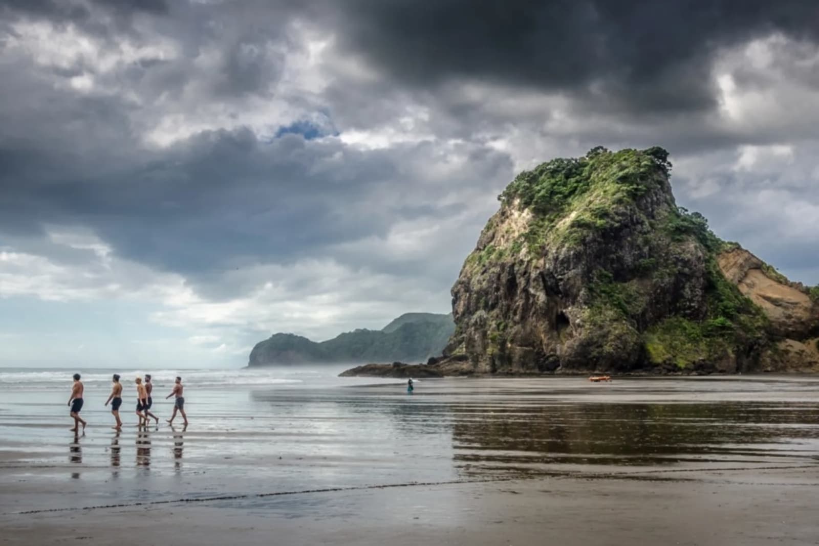 Swimmers wander across the black sands of Piha Beach at low tide with Lion Rock in the background.